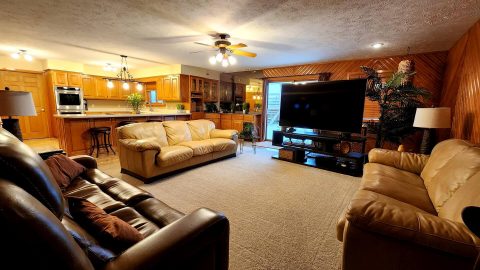 the interior of a big living room with brown and tan couches, a big screen tv, and a kitchen in the background